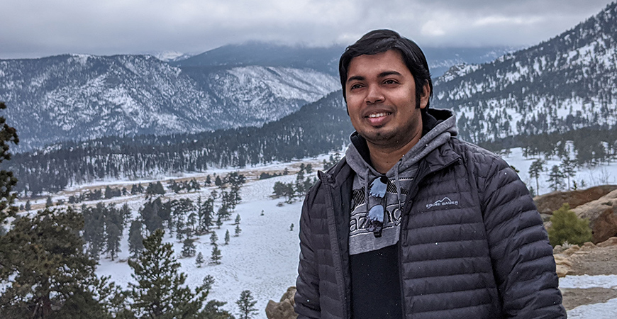 Male wearing gray jacket standing in front of snow mountains